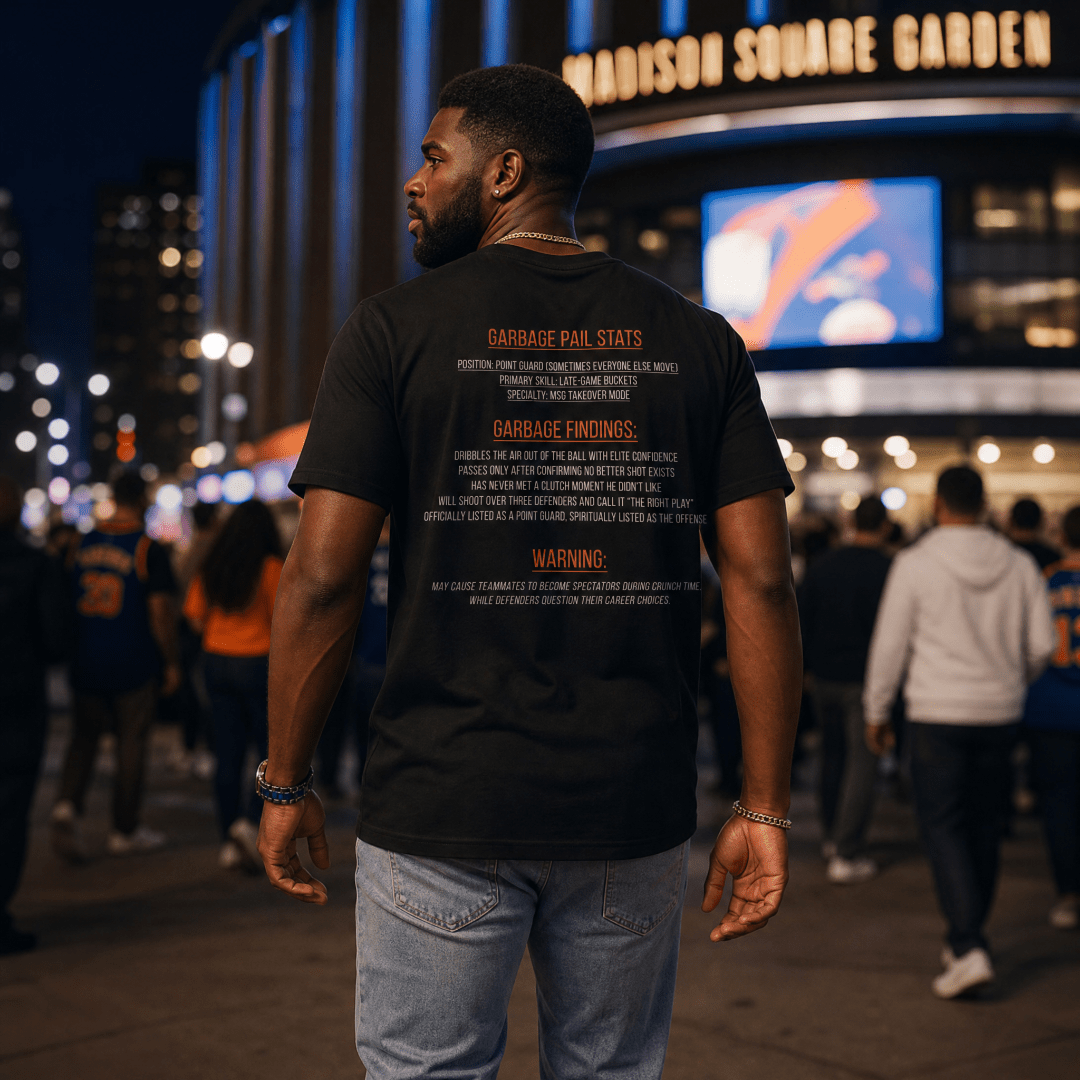 A male model walking outside Madison Square Garden at night wearing the back of a black “Captain Clutch” Garbage Pail Ballers t-shirt featuring bold orange and white New York basketball inspired typography graphics. Cinematic NYC streetwear atmosphere with realistic crowd blur and luxury handmade urban fashion style by Rebel P Customs.